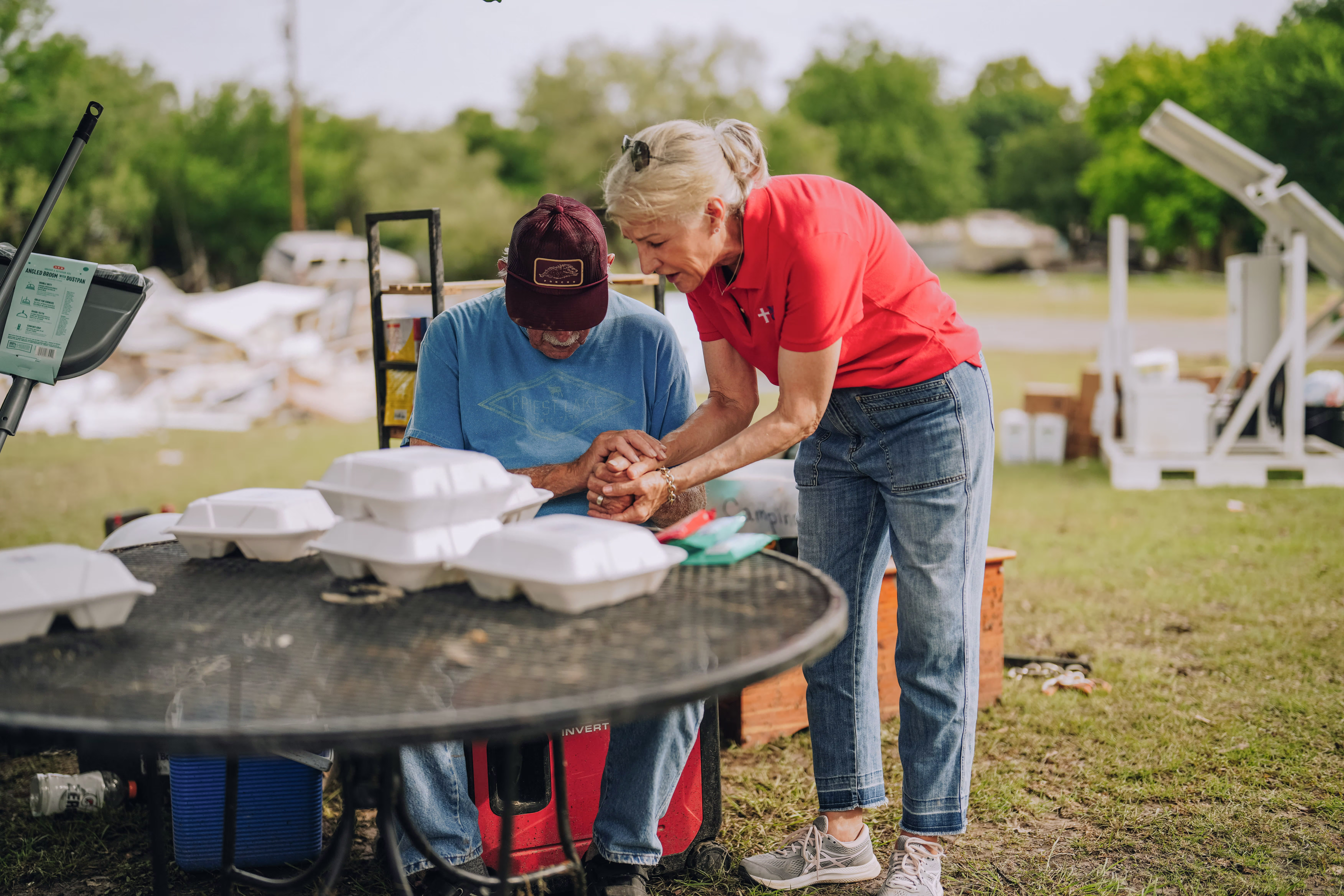 Volunteer praying with a seated man at an outdoor relief site with food containers on a table.
