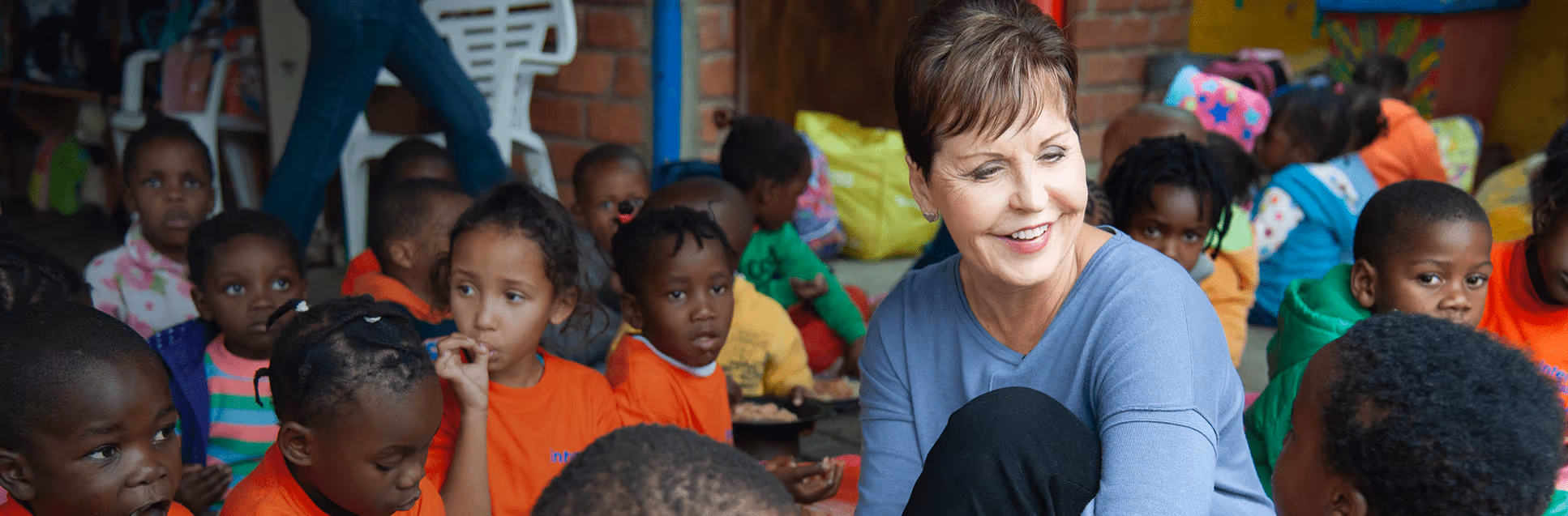 A diverse group of children sitting in a crowded room surrounding Joyce Meyer