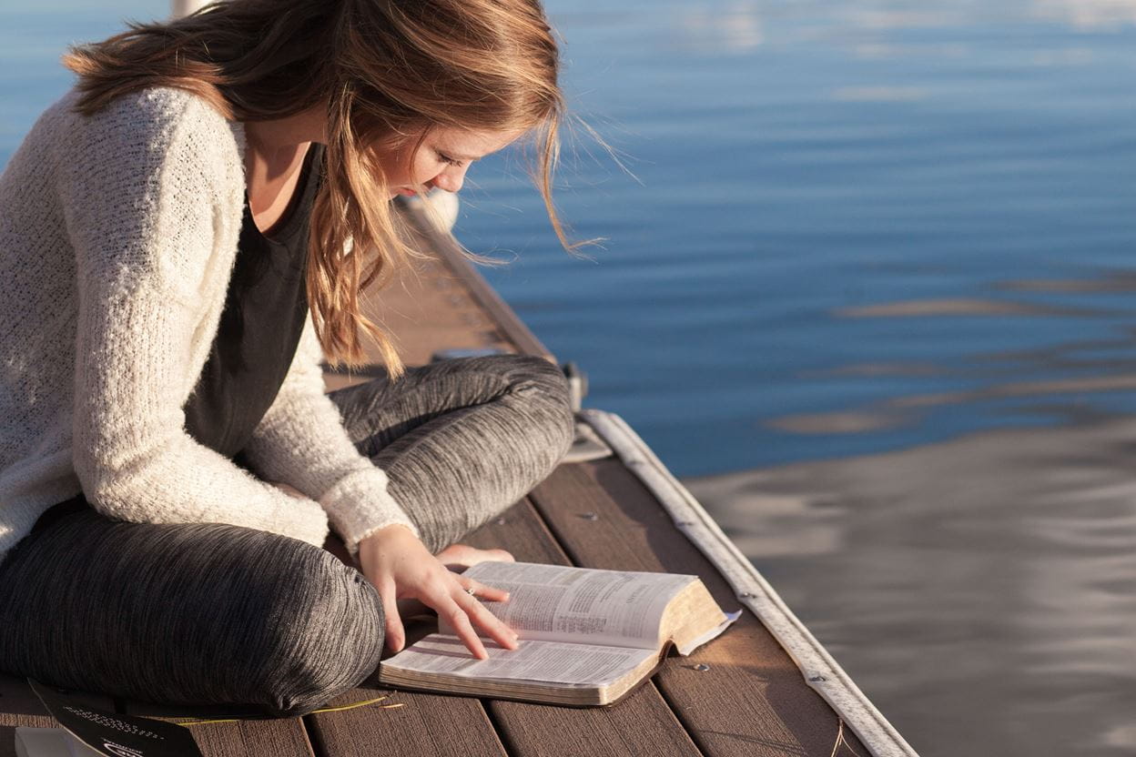 Image of a women sitting on dock reading a bible.