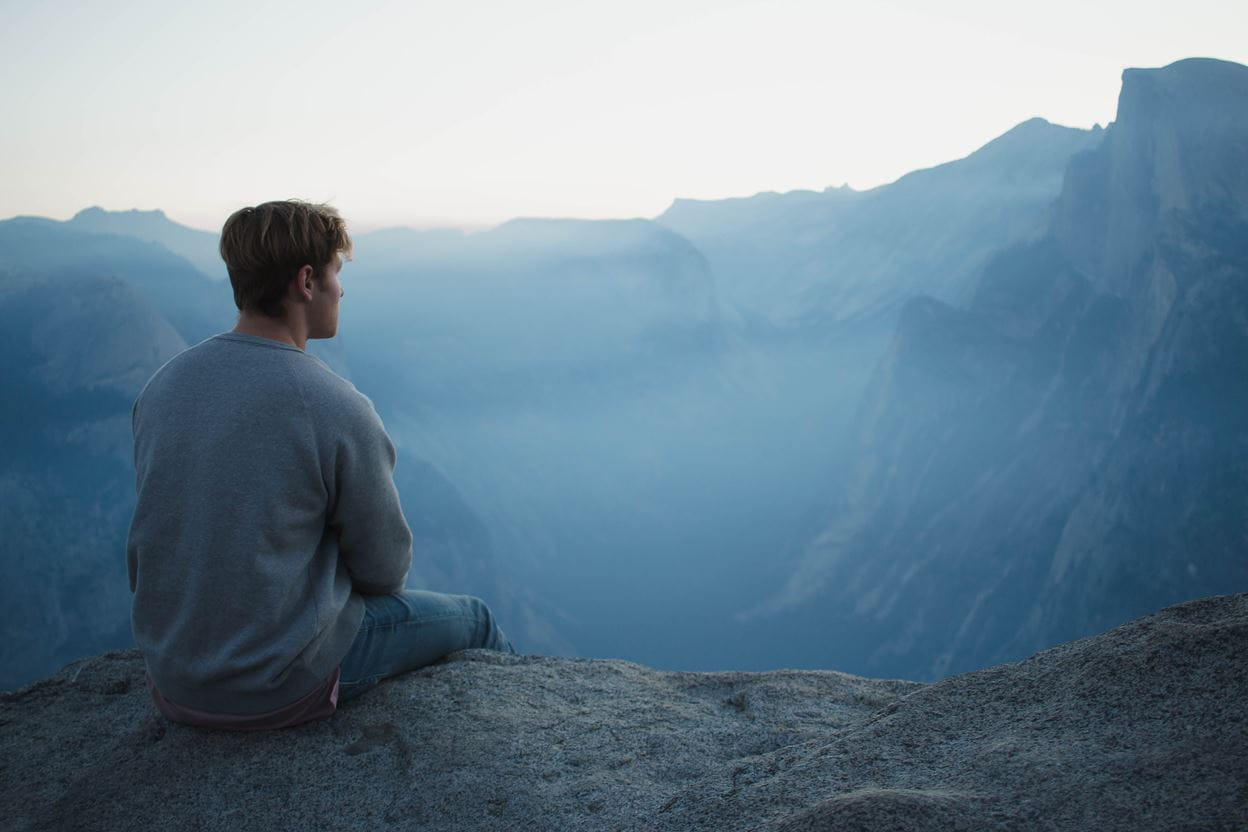 A person sits on a rocky cliff, gazing at a misty mountain landscape in the distance.