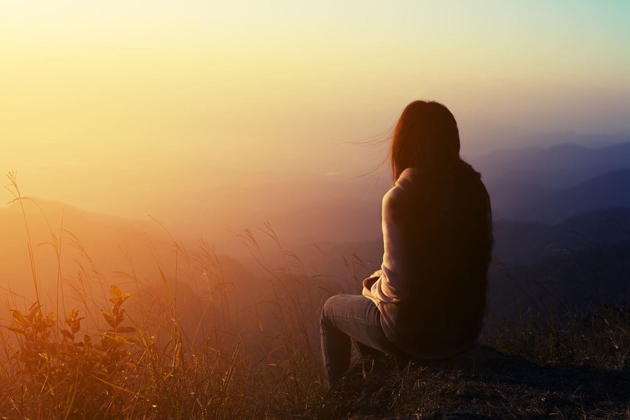 A woman, sitting on a hillside, looking out over a valley as the sun sets.