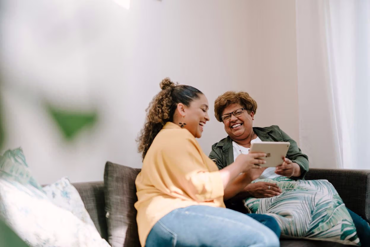 Two women sitting on a couch, looking at a tablet while smiling and laughing together.
