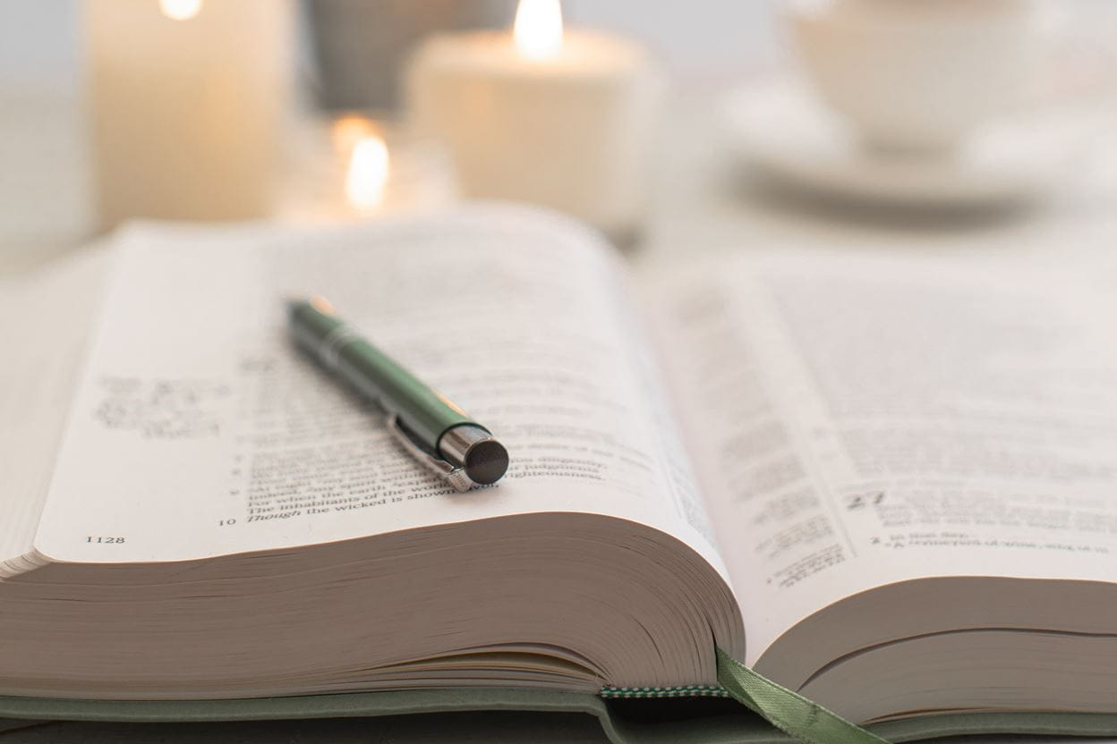 A pen sitting on an open Bible ready for reading a daily devotional with candles in the background.