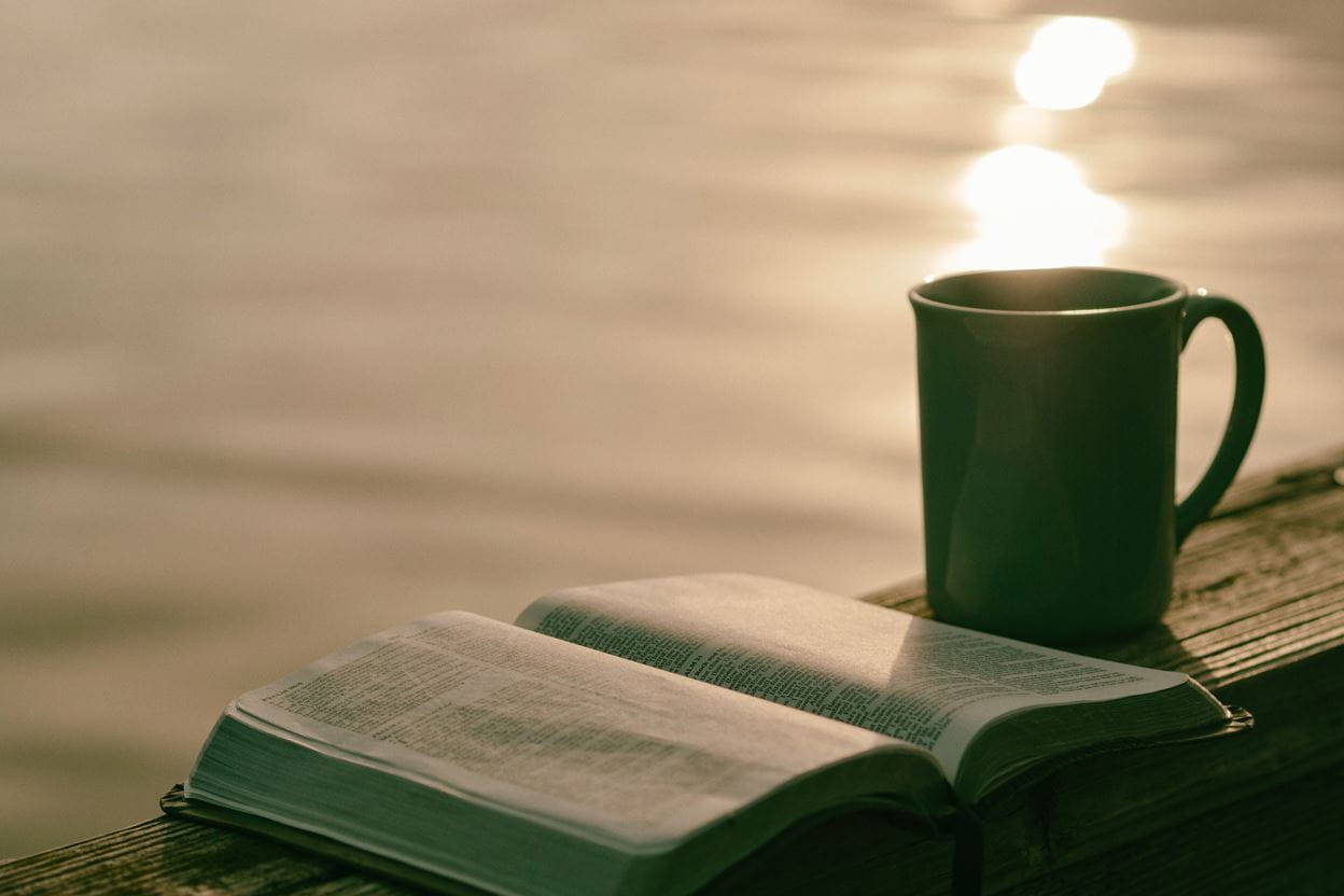 A cup of coffee and an open Bible ready for reading a daily devotional sitting by a lake.