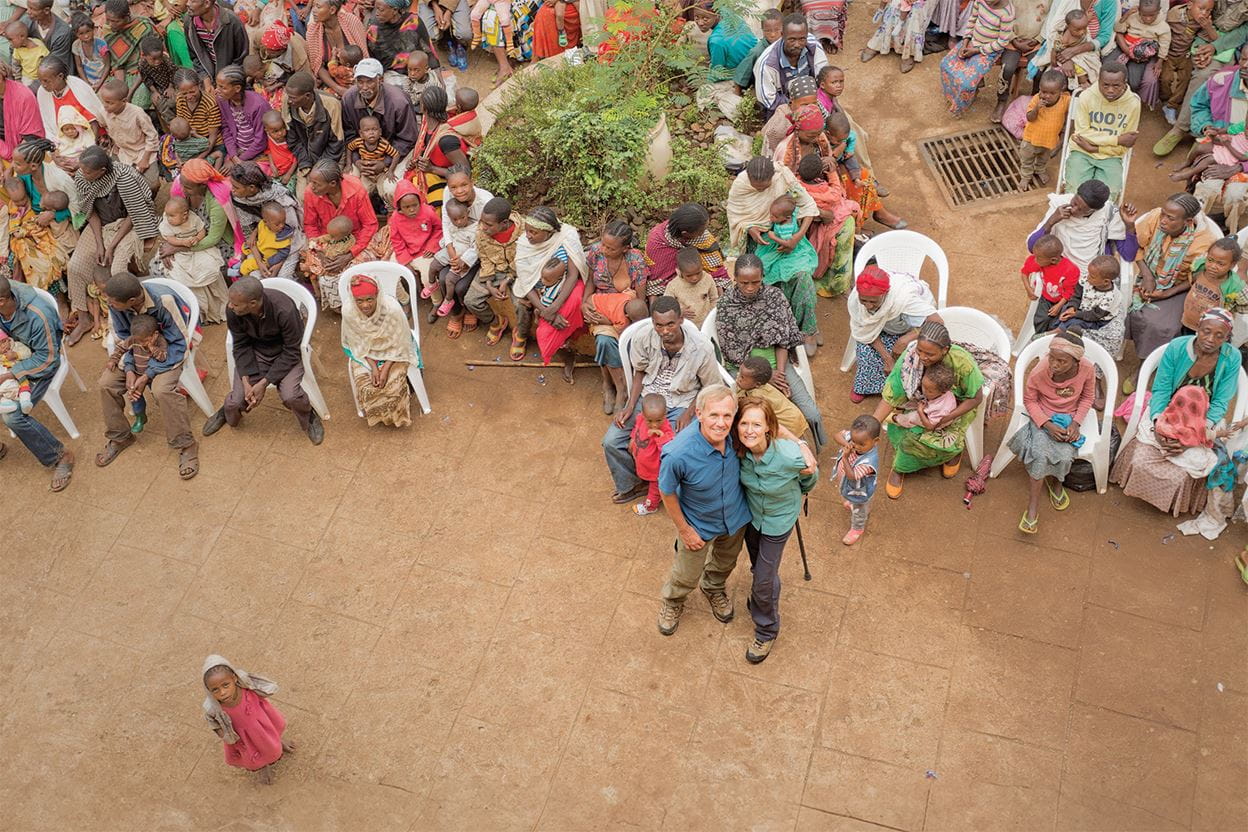 A couple stands among a large seated crowd outdoors, looking up and smiling.