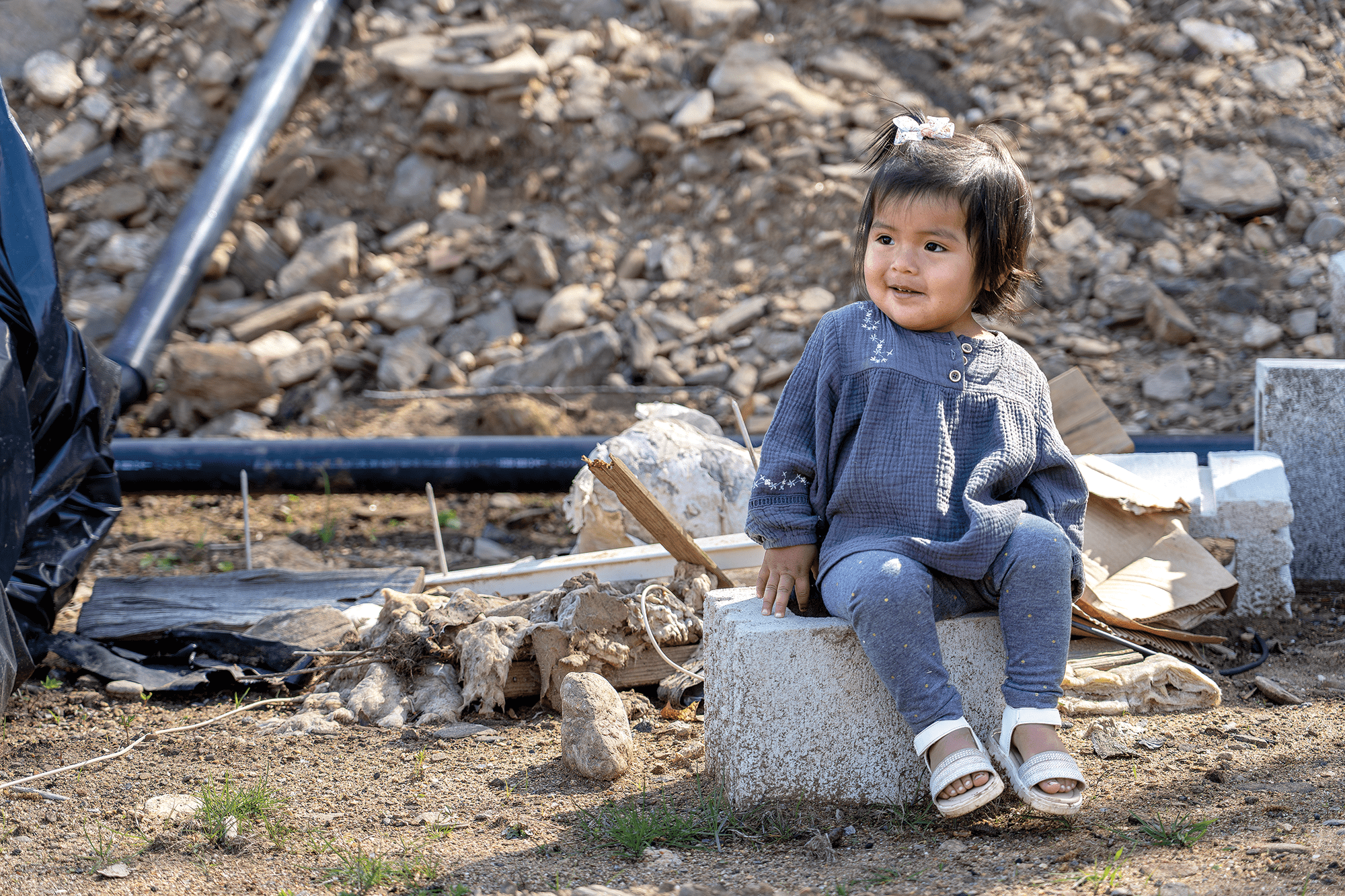 Young child sitting on a concrete block amid rubble and debris.