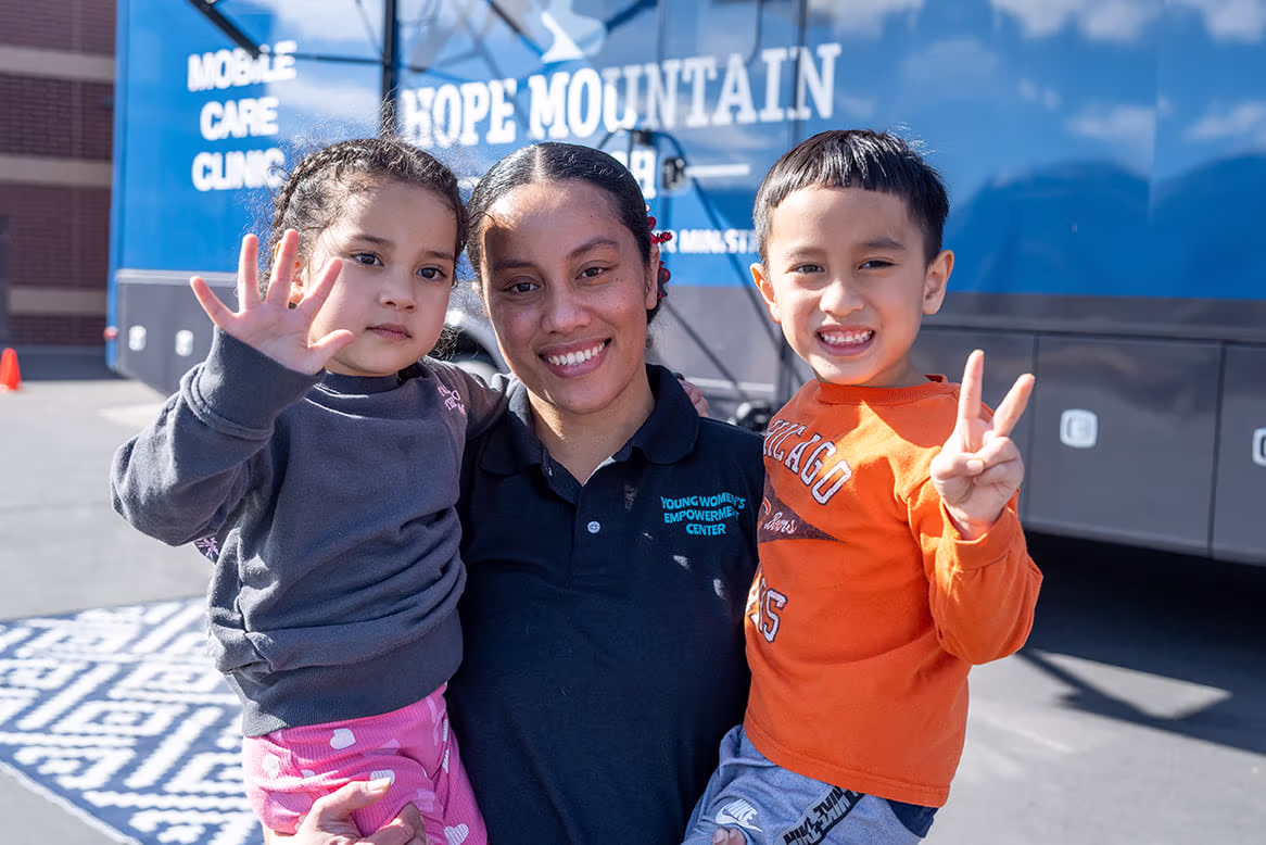 A woman and two young children smiling in front of the Hope Mountain Mobile Care Clinic.